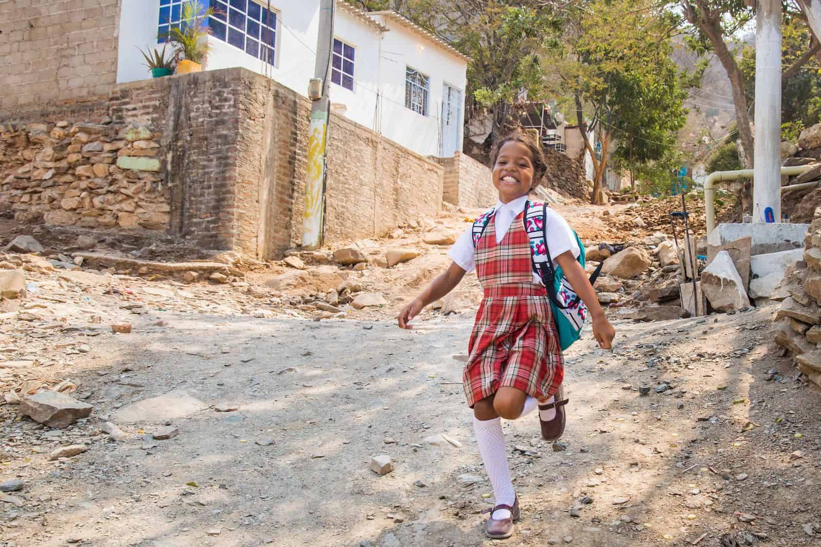 A girl in a red plaid dress wearing a backpack skips down a dirt road with a broad smile on her face.