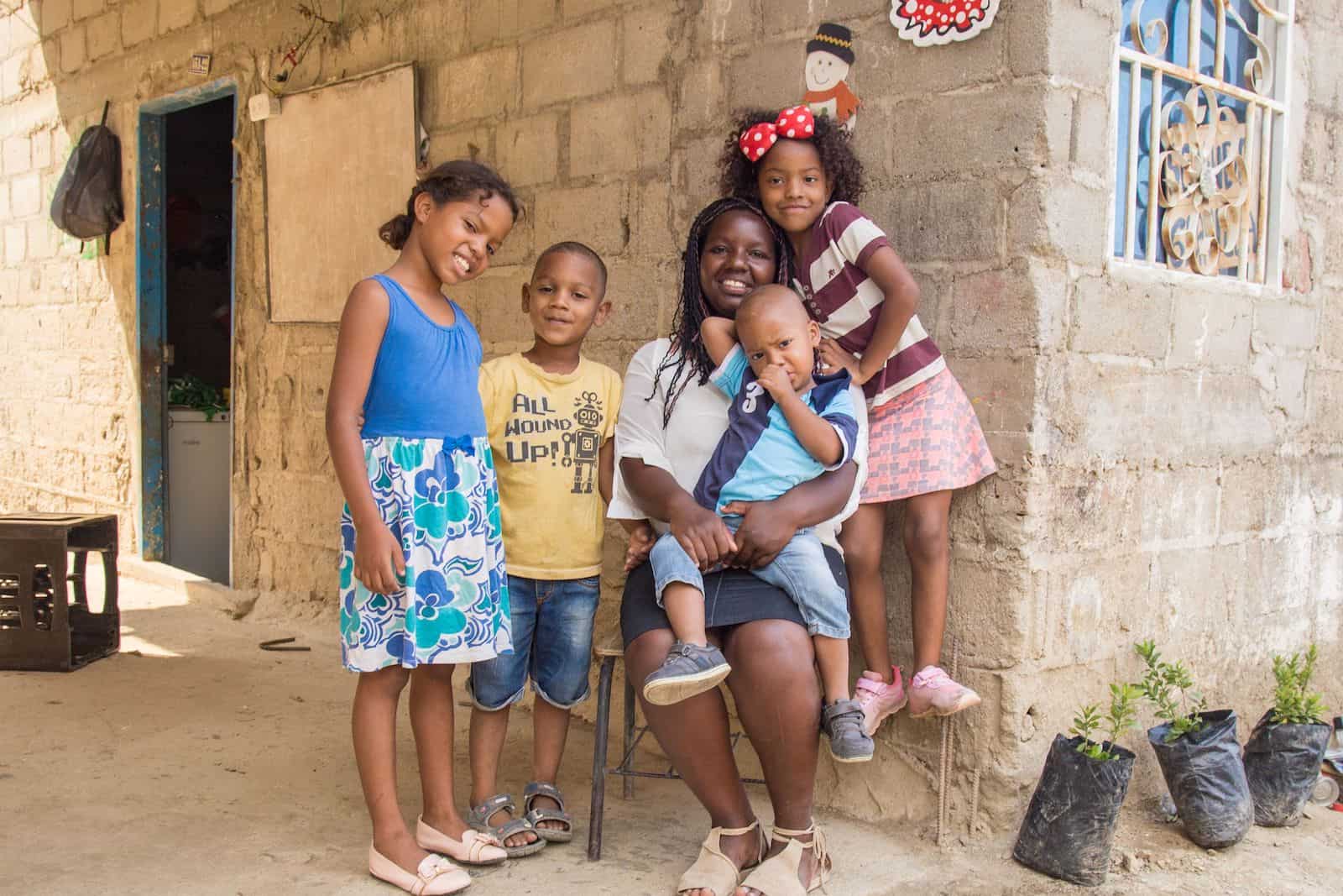 A woman sits in a chair with a young boy in his lap, surrounded by two girls and a boy in front of a cement brick home. 