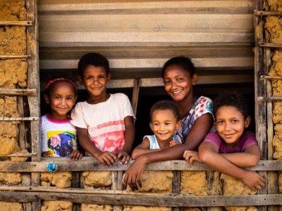 A woman stands at a windowsill of a mud and wood home, surrounded by four children.