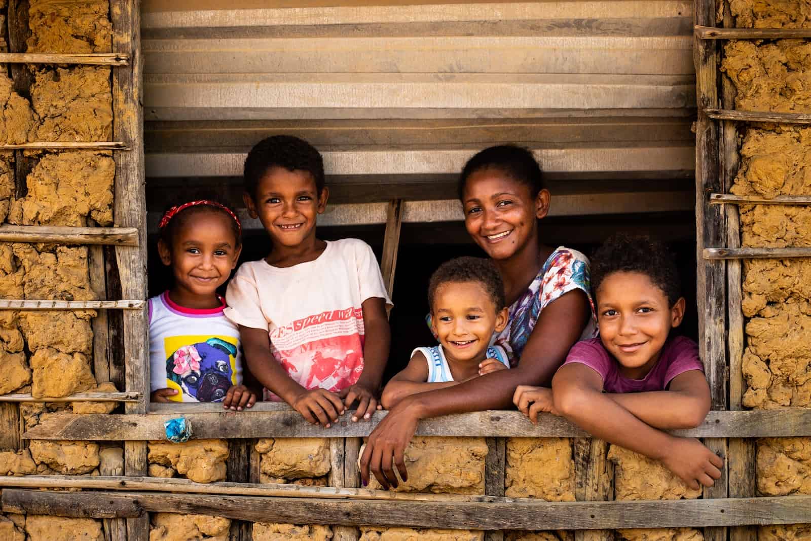 A woman stands at a windowsill of a mud and wood home, surrounded by four children.