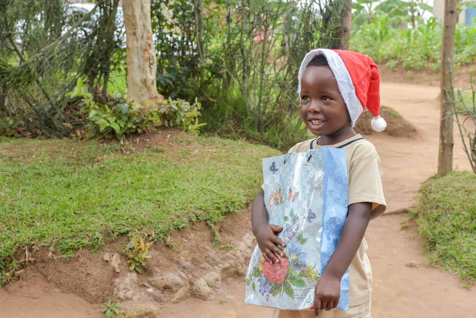 A boy in a tan shirt and Santa cap holds a gift bag, smiling and standing on a dirt path.