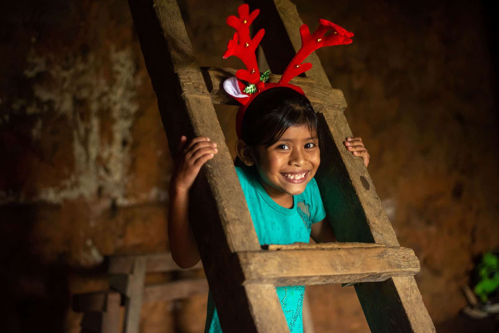 A girl wearing a green shirt and reindeer horns peeks out from a wooden ladder. 