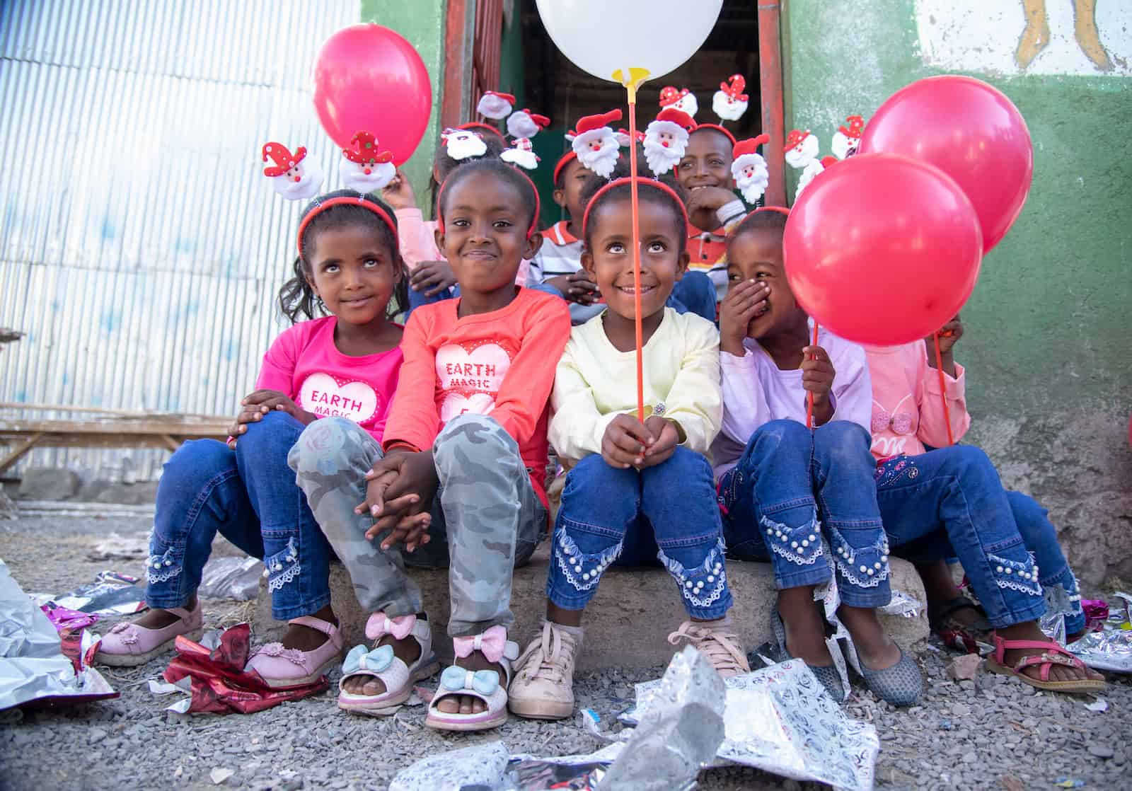 A group of children sit on a church stoop, holding balloons and wearing Santa Claus headbands.
