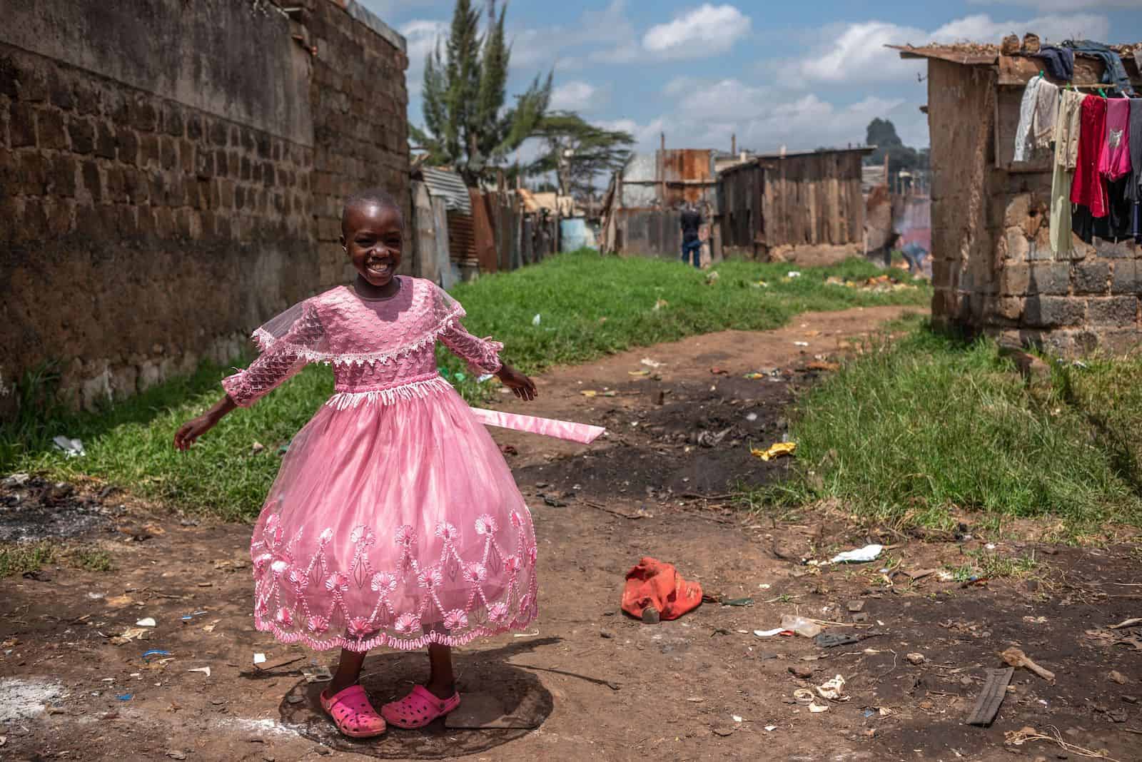 A girl spins in a pink dress, standing on a dirt path in front of a shanty home with laundry hanging on a line.