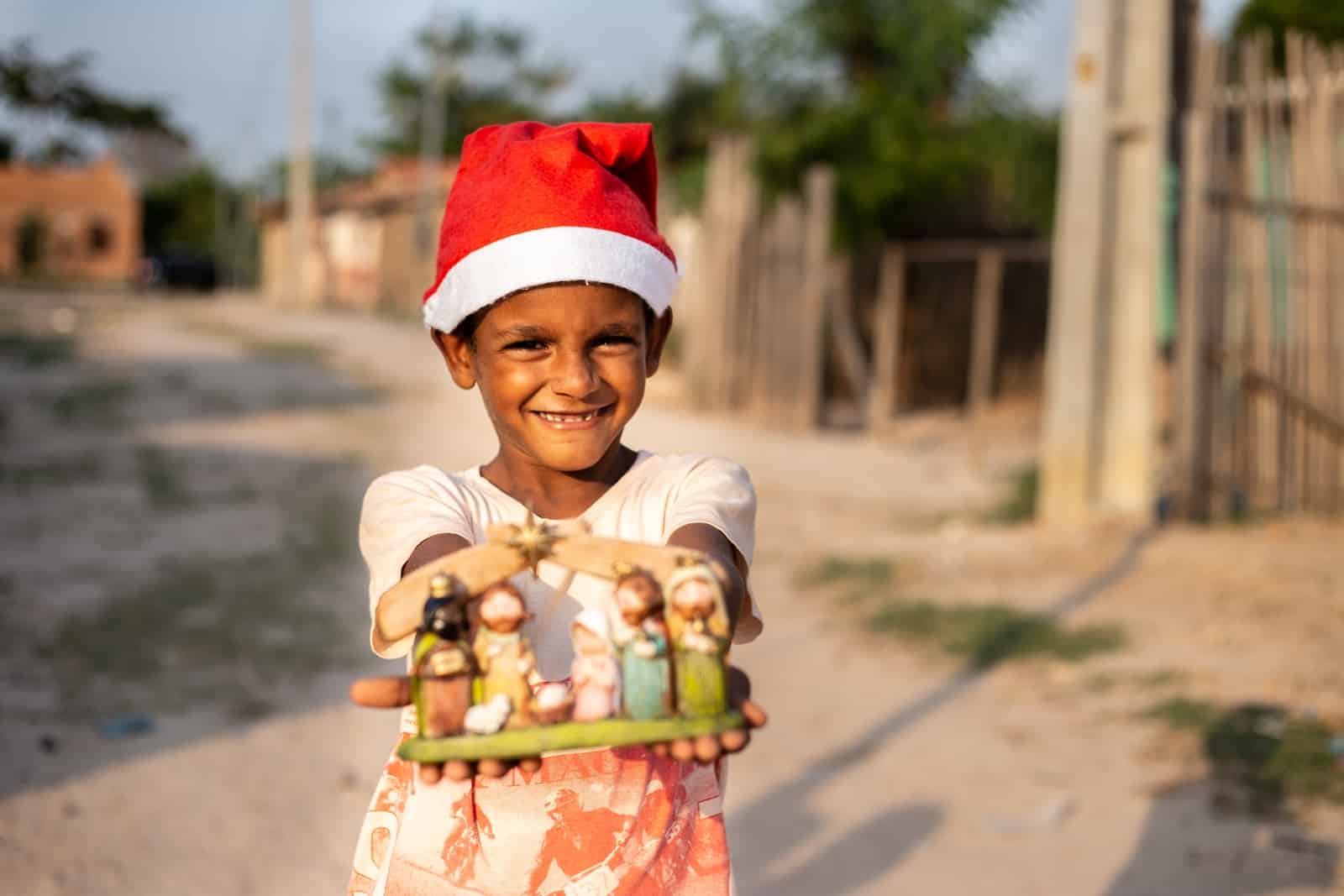 A boy wearing a red Santa cap holds out a small nativity set. 