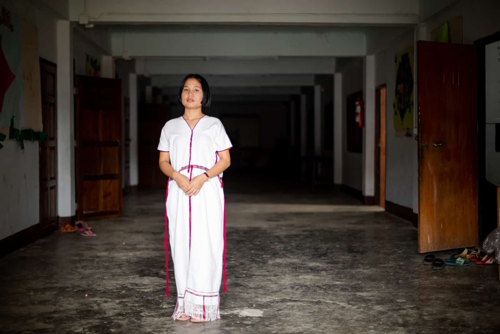 A girl in a long white dress stands in a long dark hallway.