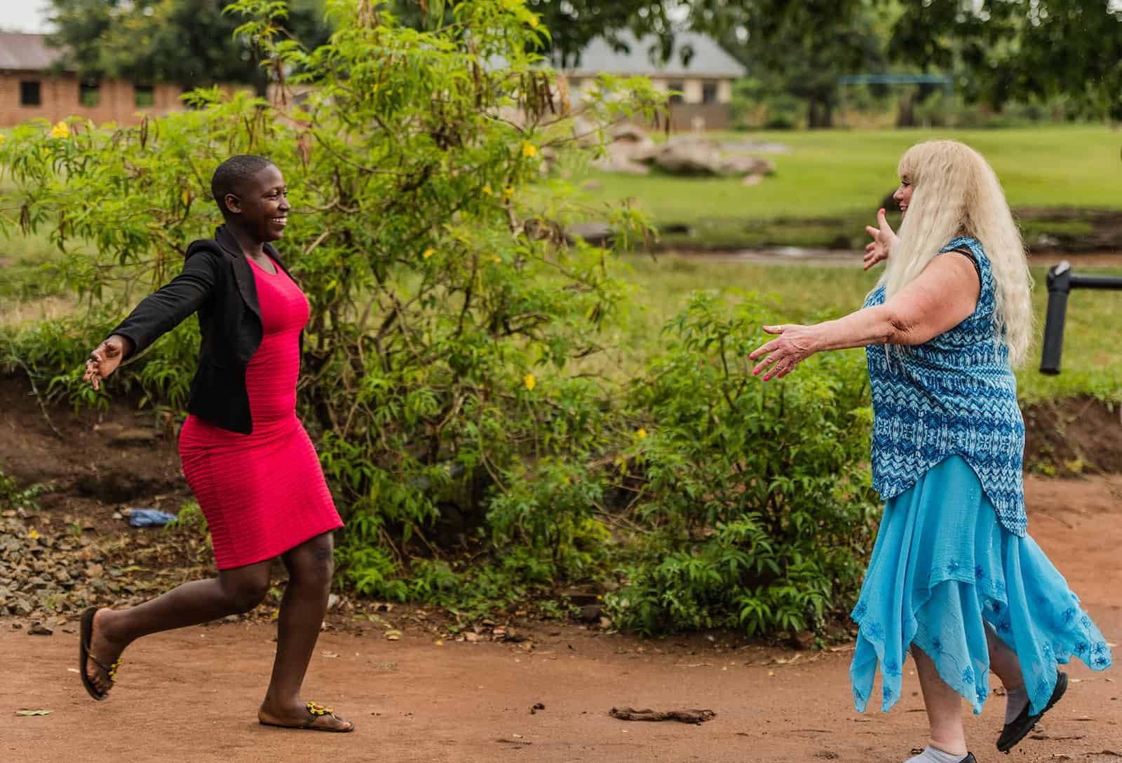 A girl in a red dress runs toward a woman in a blue dress with outstretched arms.