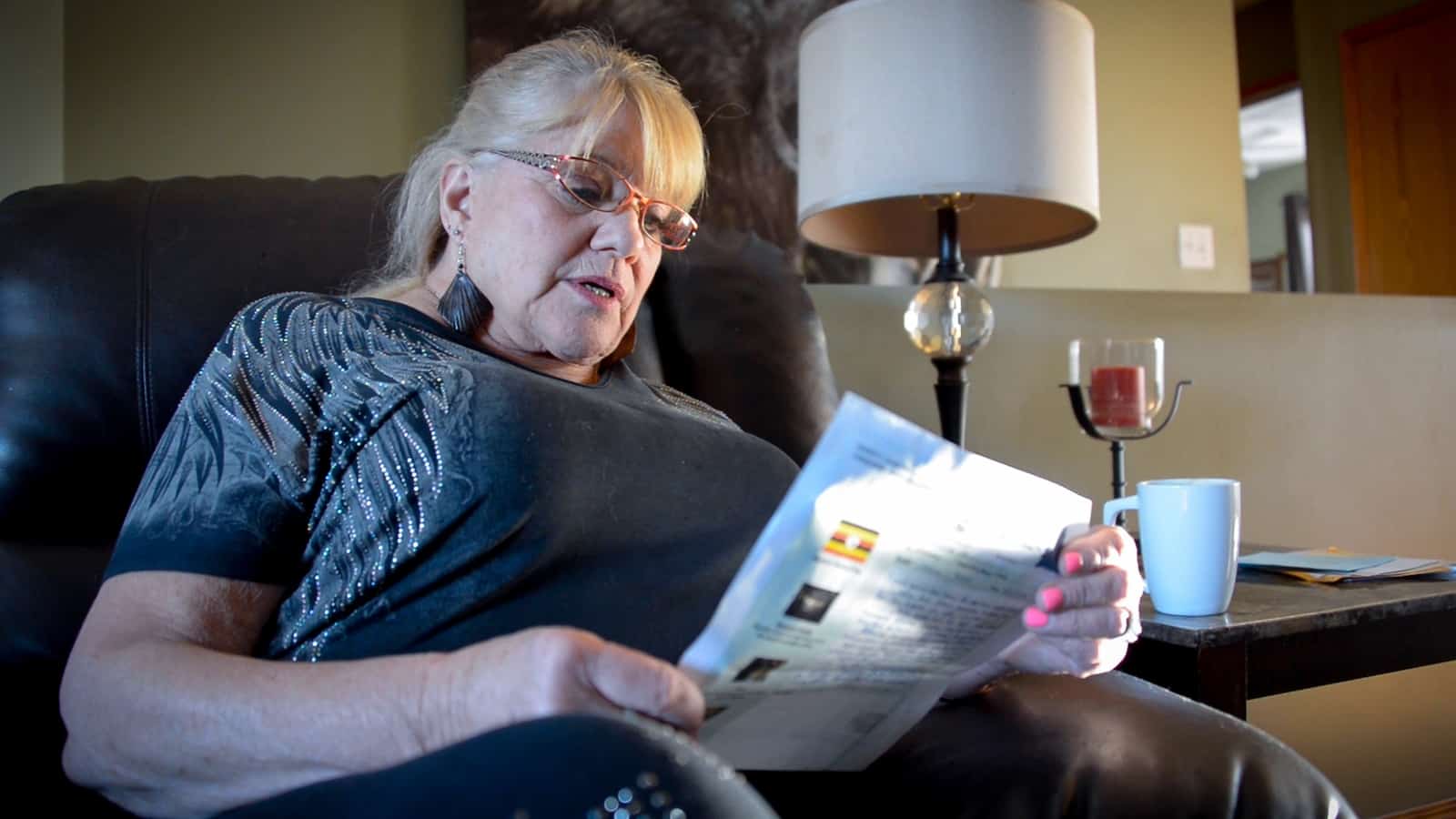 A woman in a black shirt sits inside in a chair reading a letter. 