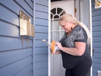 A woman in a black shirt checks the mail out of a box on the front porch of a house.