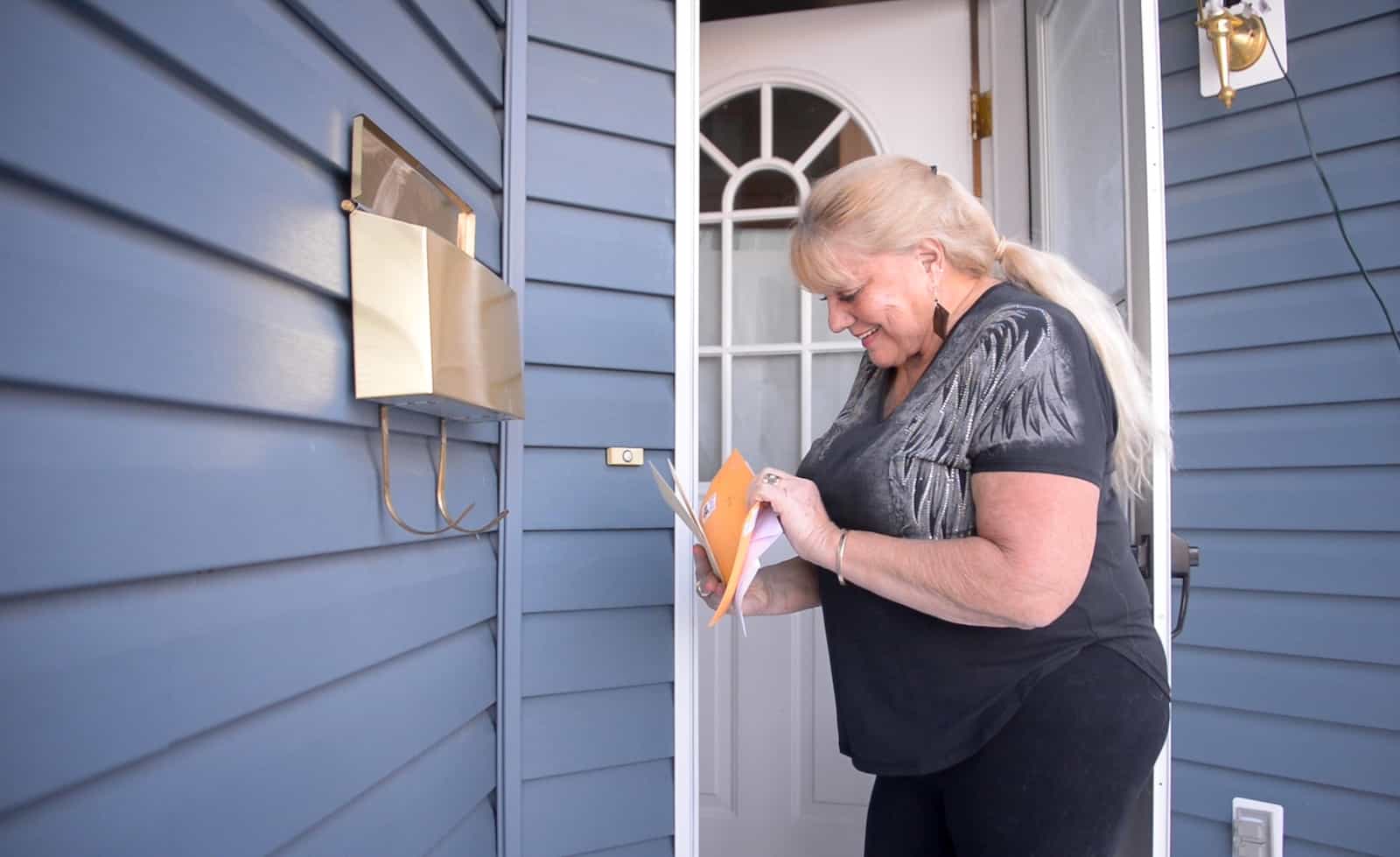 A woman in a black shirt checks the mail out of a box on the front porch of a house. 