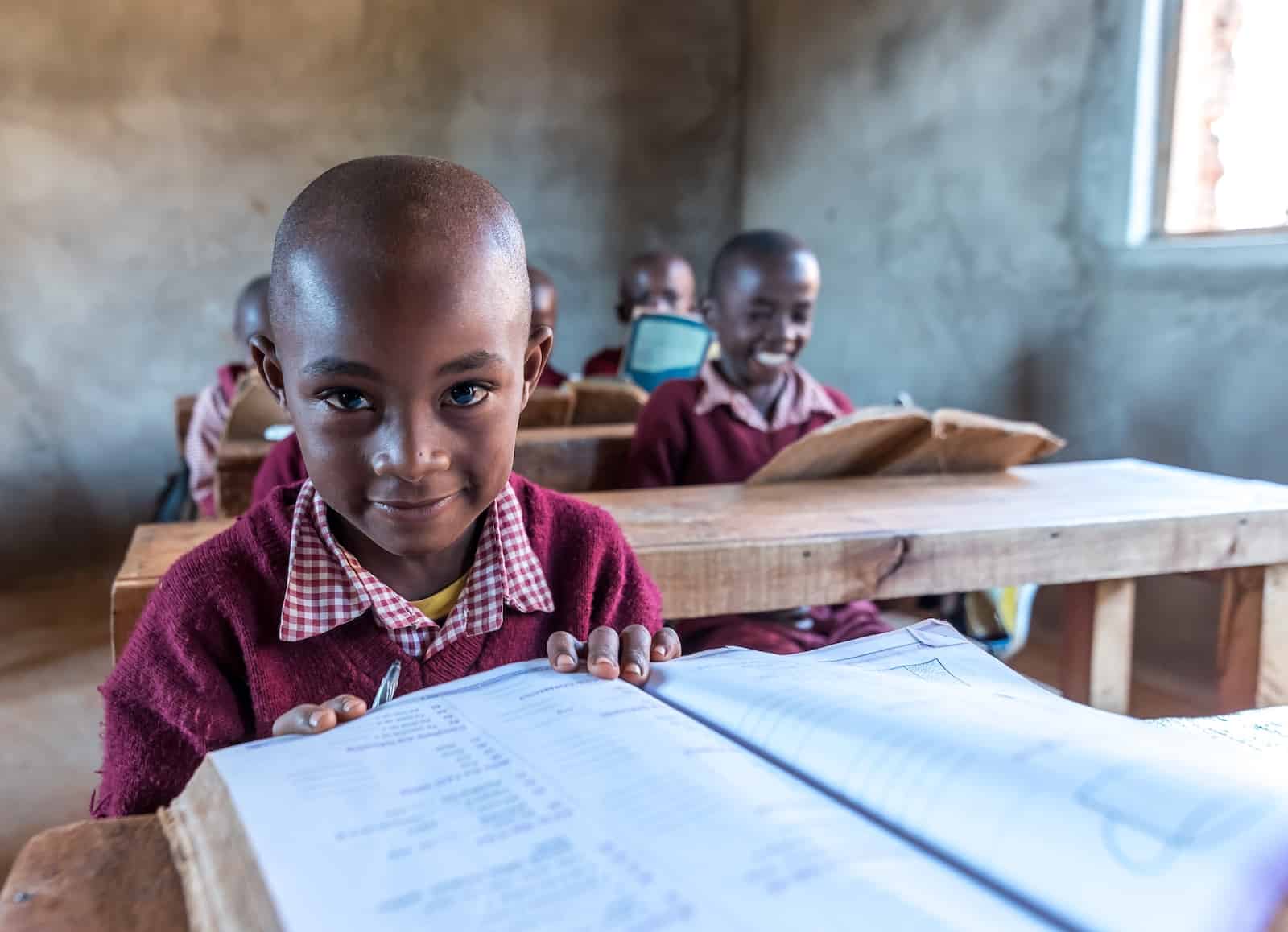 A boy in a red sweater sits at a desk in a classroom, reading a book.