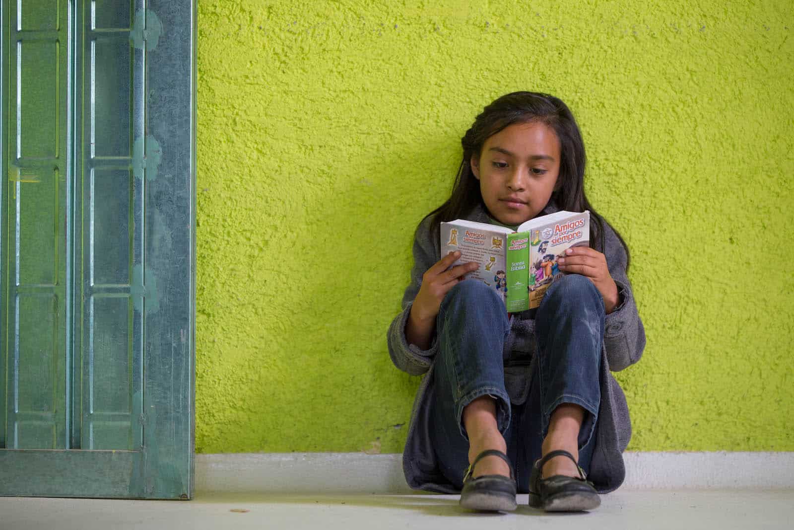 A girl sits against a lime green wall, reading a book. 