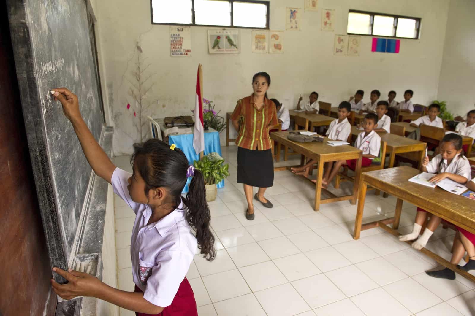 A girl stands at a chalkboard, writing, while children sit at desks watching.