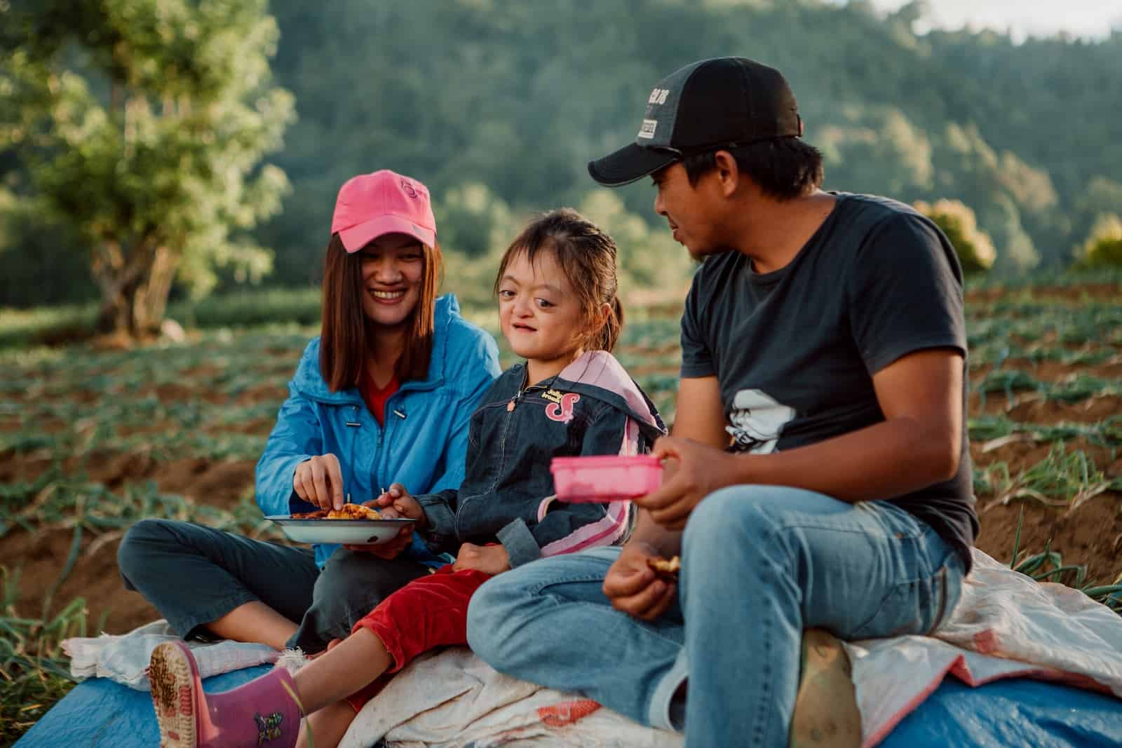 A girl sitting on the ground surrounded by her parents holding food containers with greenery in the background.