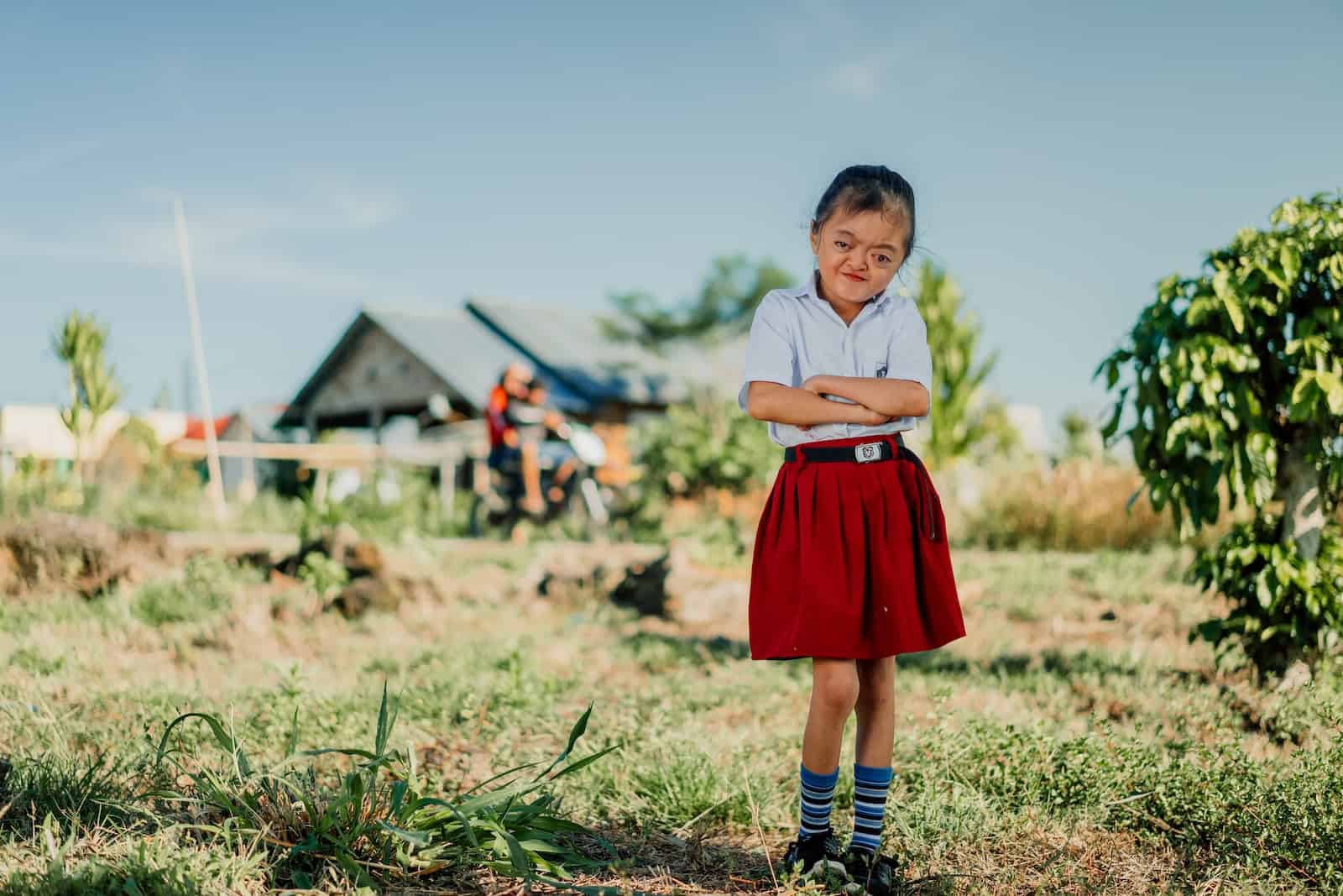 A photo of a girl with Apert Syndrome in a red and white school uniform stands in front of a house, with her arms crossed in front of her.