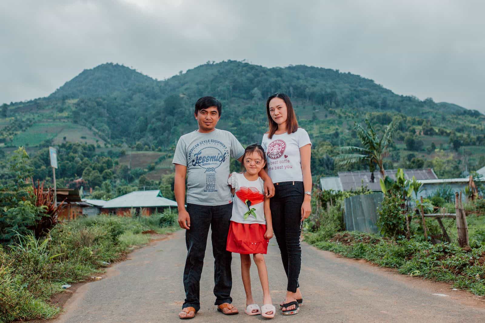 A photo of a girl with Apert Syndrome standing with two adults on a road in front of a mountain.