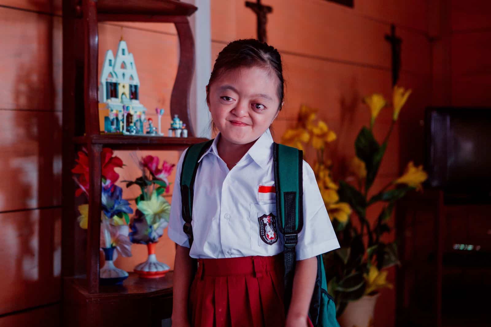 A photo of a girl with Apert Syndrome wearing a red and white school uniform and backpack, standing inside against a red wall.