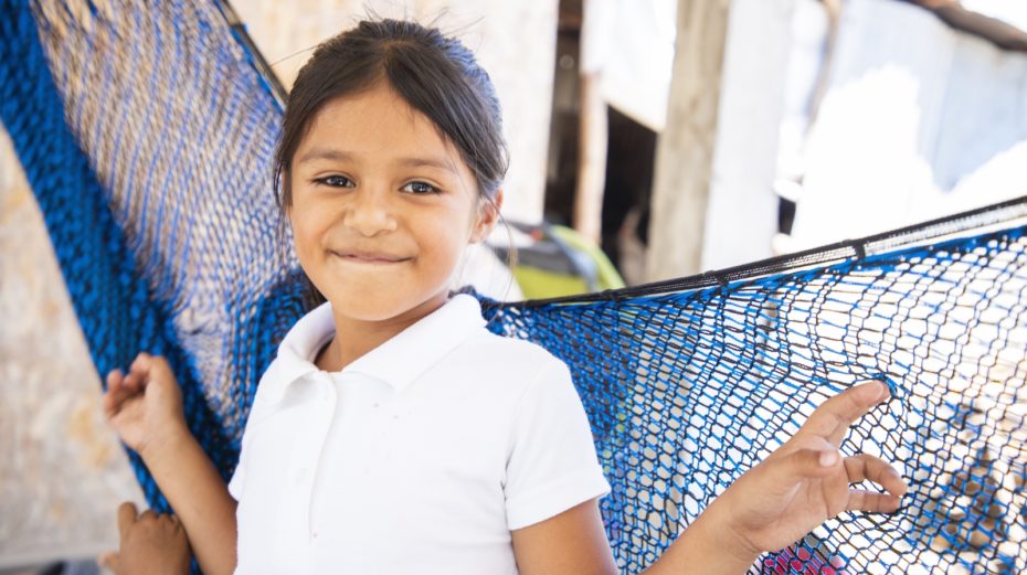 A girl in a white shirt stands in front of a blue hammock, smiling.