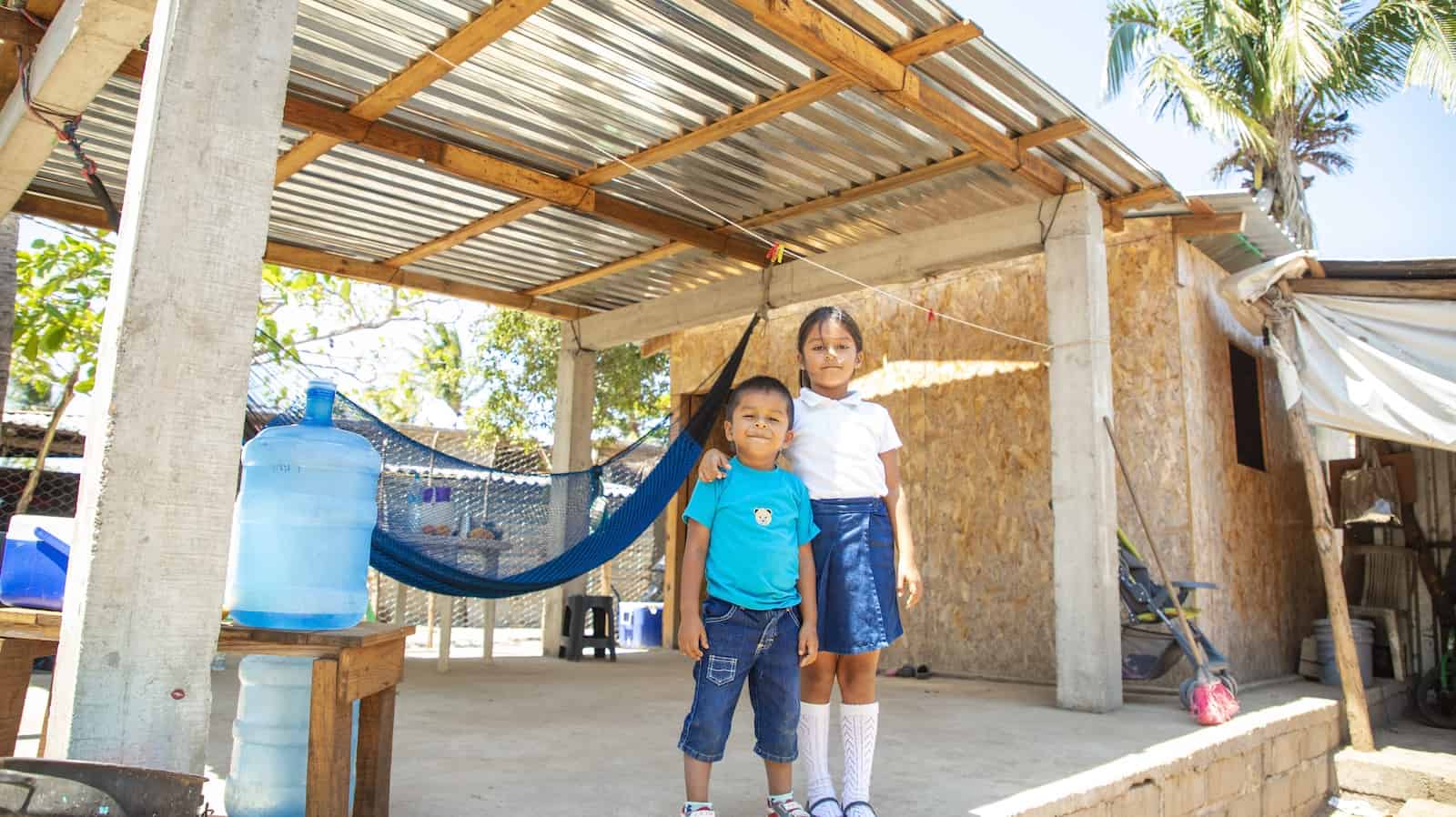 Two children stand in front of a home built after the Mexico earthquake made from plywood with a tin roof shelter in front of it. 
