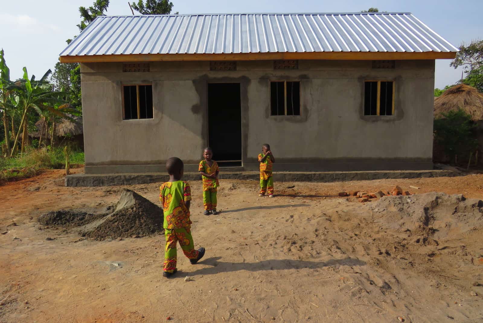 Three small children in orange and green patterned outfits stand in front of a concrete home with a metal roof, surrounded by thatched roof homes.