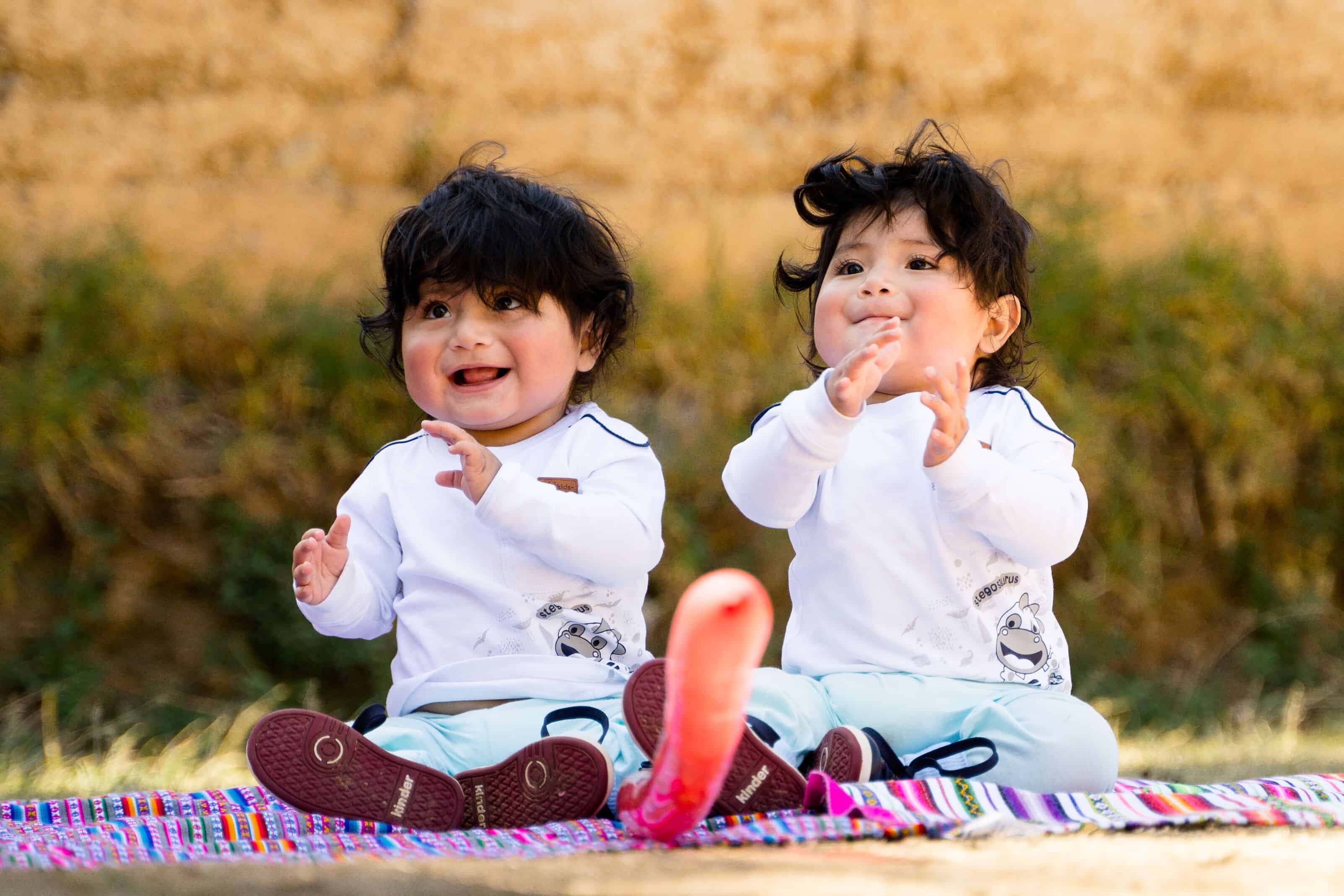 Two cute babies sit on a blanket, smiling
