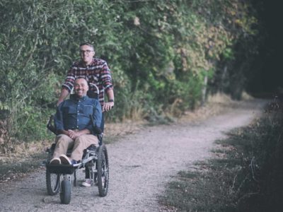One man stands behind another man sitting in a wheelchair on a dirt path.