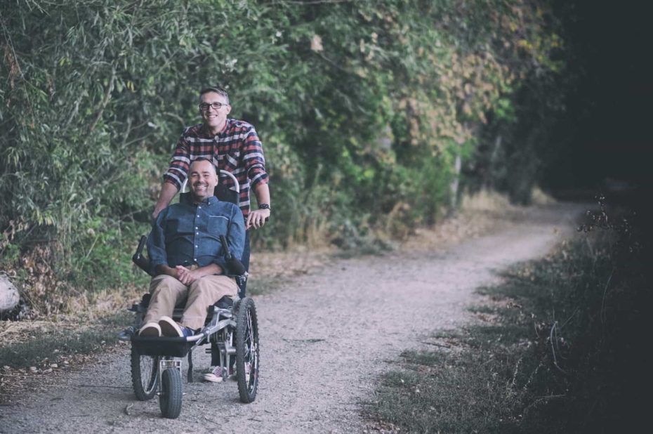 One man stands behind another man sitting in a wheelchair on a dirt path.