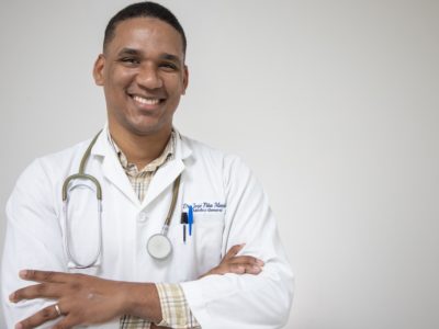 A man wearing a white doctor's coat and stethoscope around his neck smiles, arms crossed.