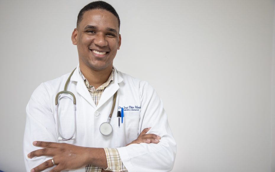 A man wearing a white doctor's coat and stethoscope around his neck smiles, arms crossed.