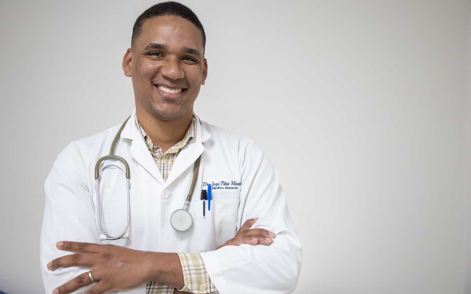 A man wearing a white doctor's coat and stethoscope around his neck smiles, arms crossed. 