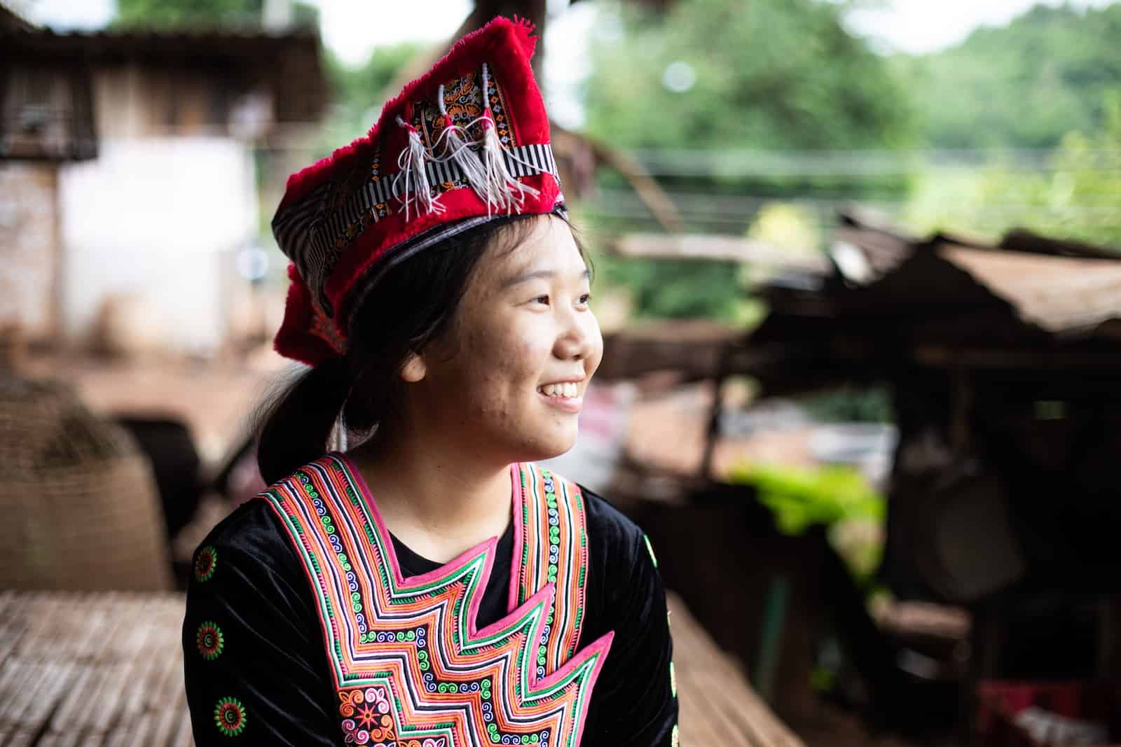 A girl wearing traditional Hmong clothing looks to the side, smiling.