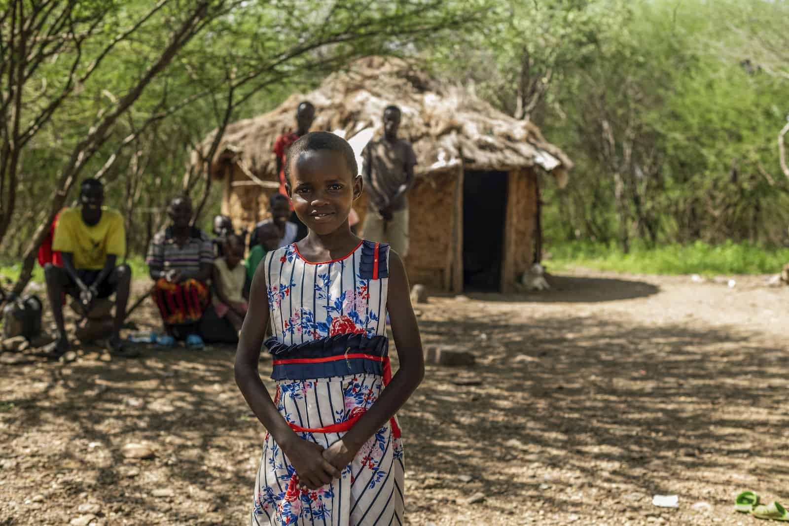 A young girl stands in front of a grass thatched hut with her family sitting behind her.