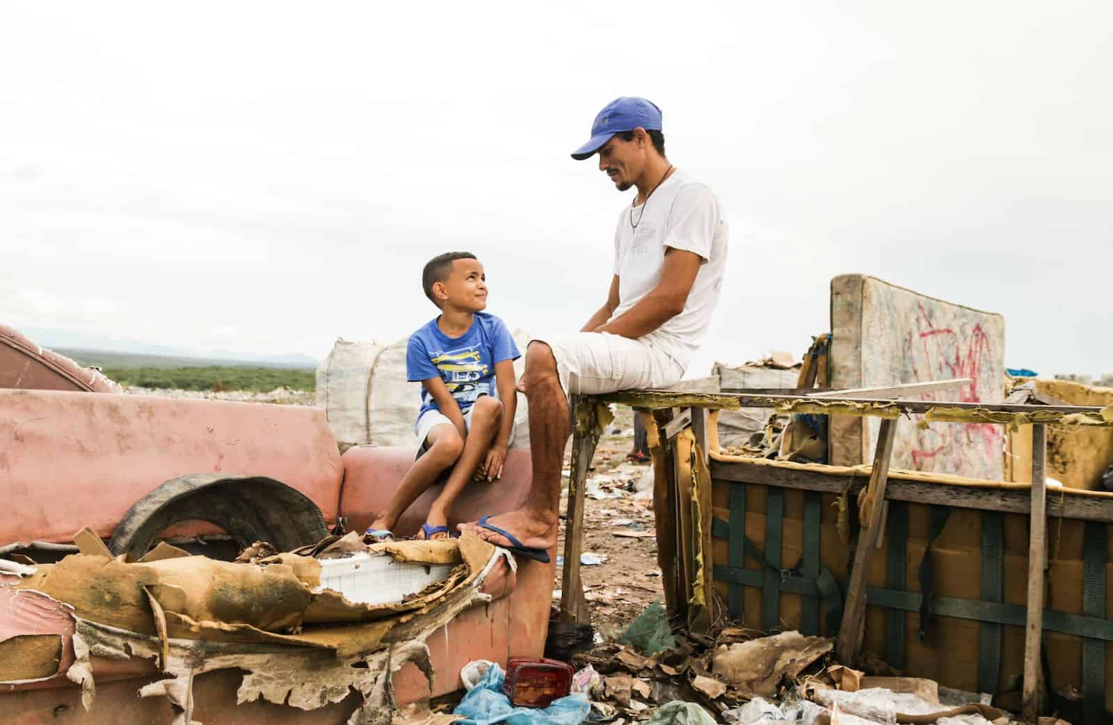 A man and son sit on old furniture in a landfill, looking at each other.