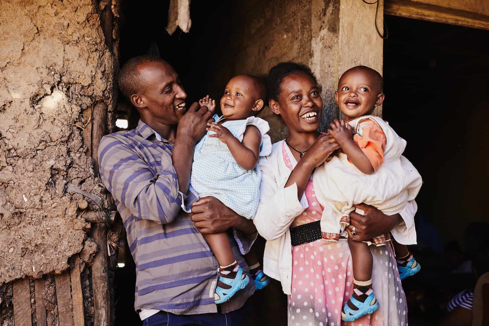 A man and woman stand outside a mud home holding twin babies.