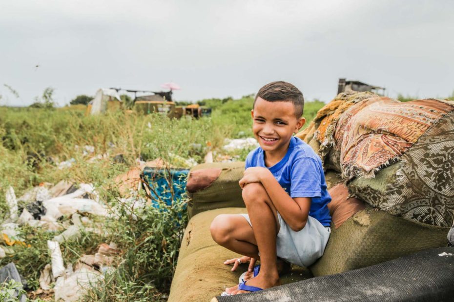 A boy sits on an old sofa in a garbage dump.