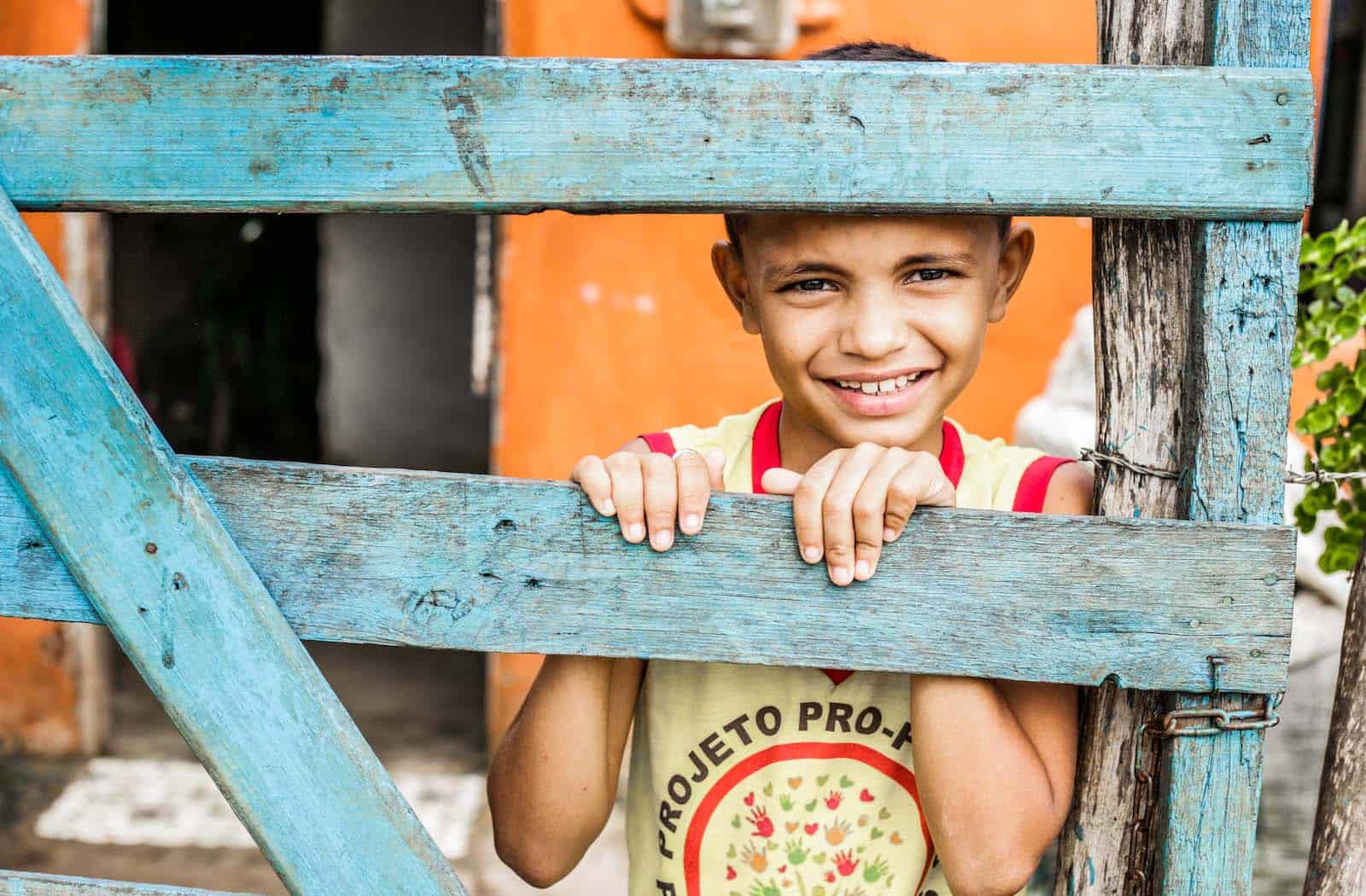 A boy stands behind a green fence, peeking through the slats. 