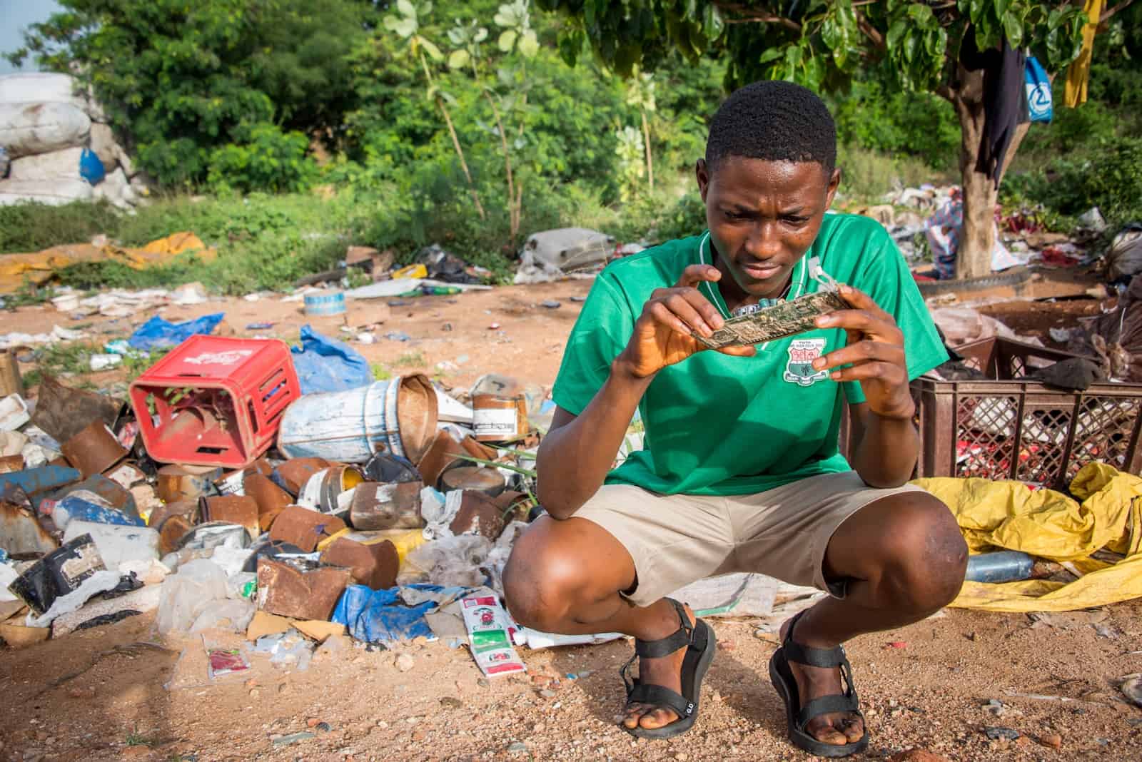 A young man squats surrounded by garbage, looking at a circuit board.