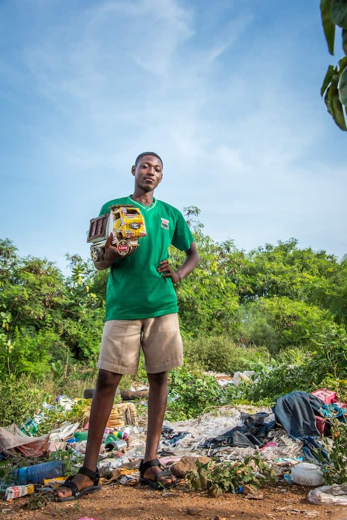 A young man stands surrounded by garbage, holding a handmade toy truck.
