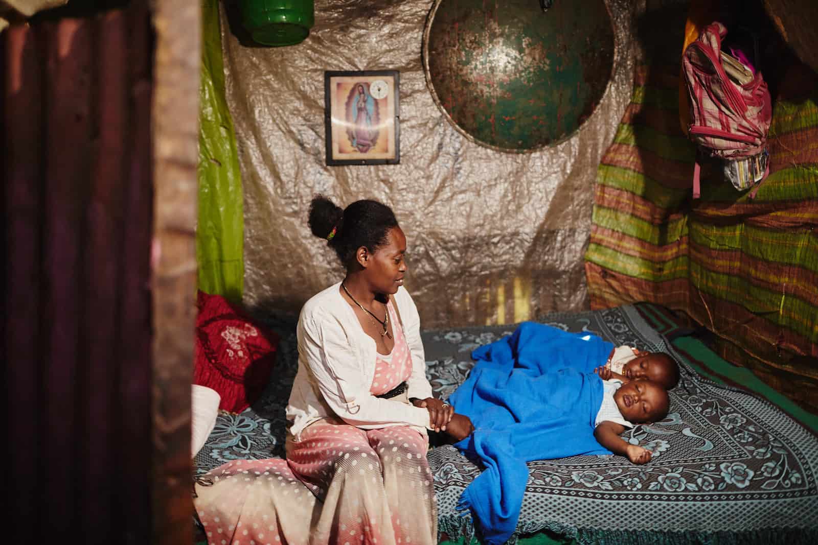 A woman sits on a bed looking at two sleeping babies. 