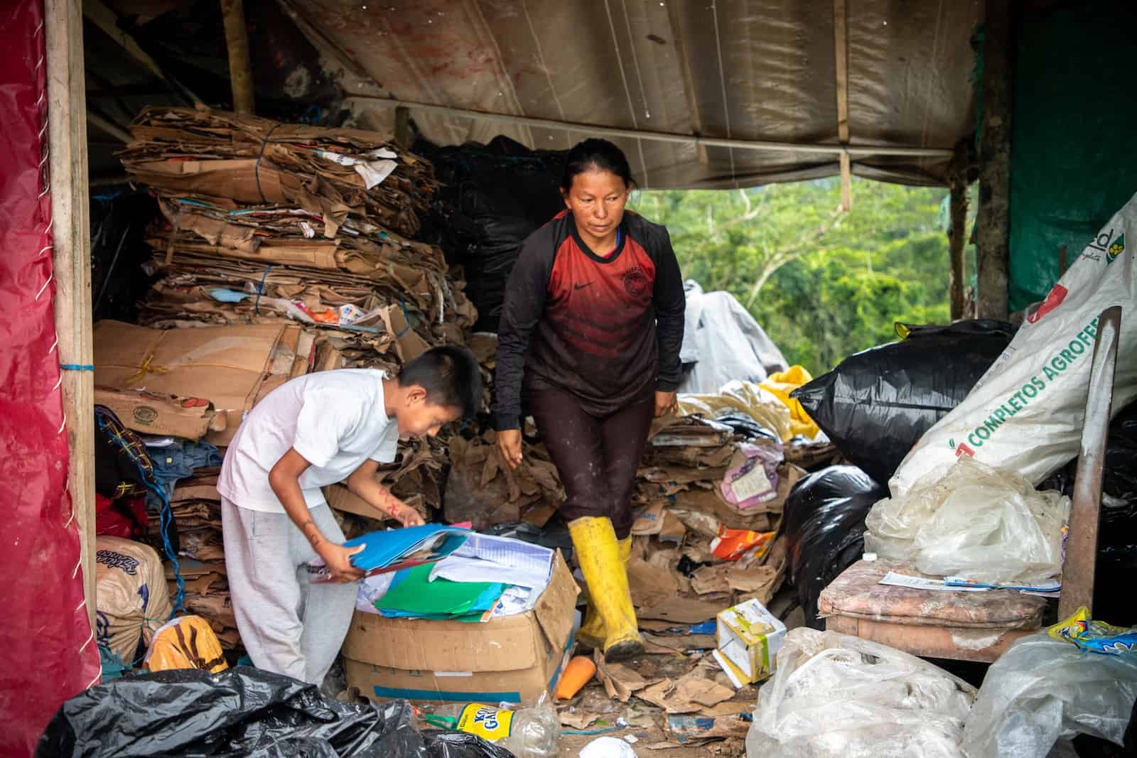 A woman and boy stand in a landfill warehouse, sorting cardboard.