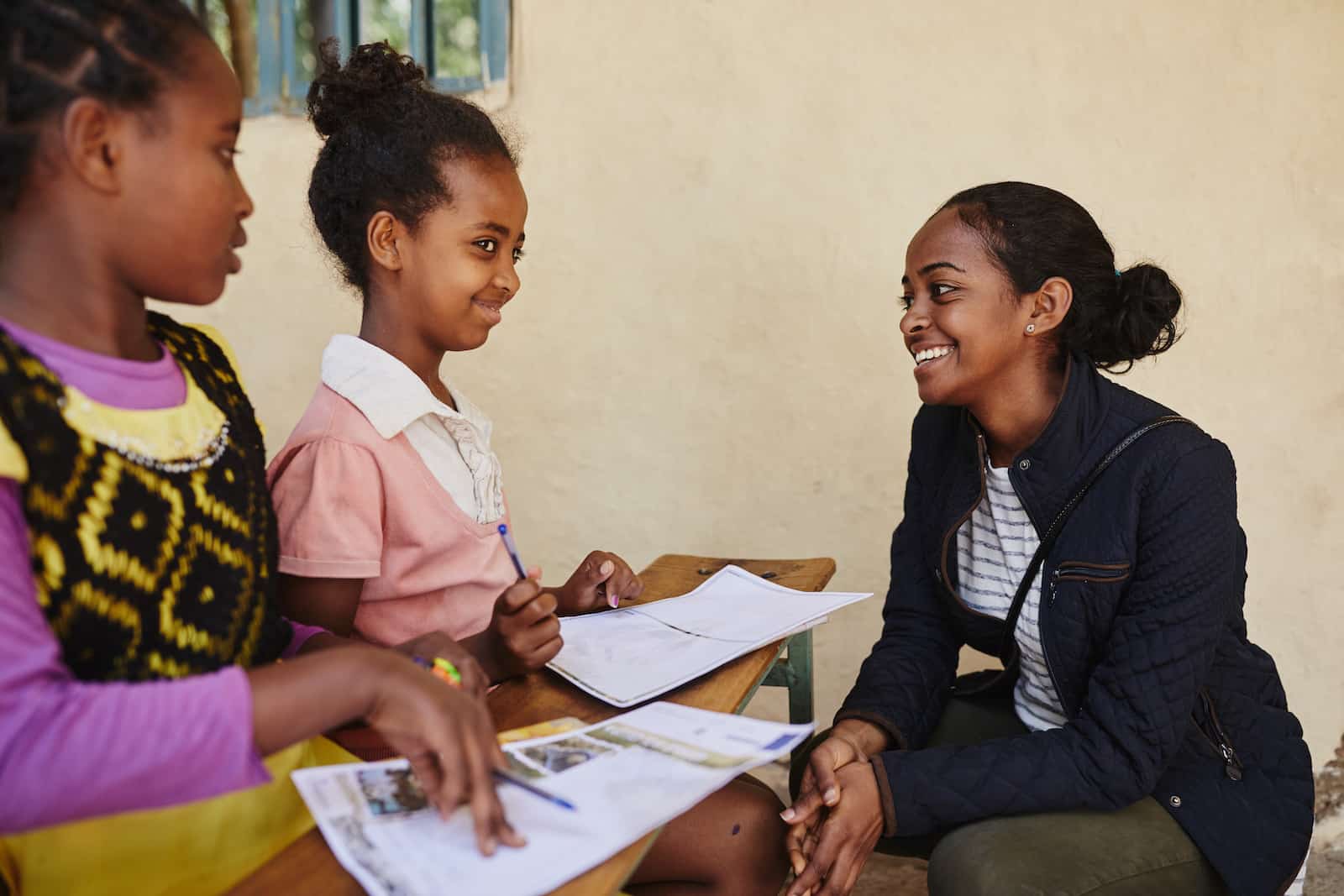 A letter translator kneels in front of two girls at a desk writing letters.