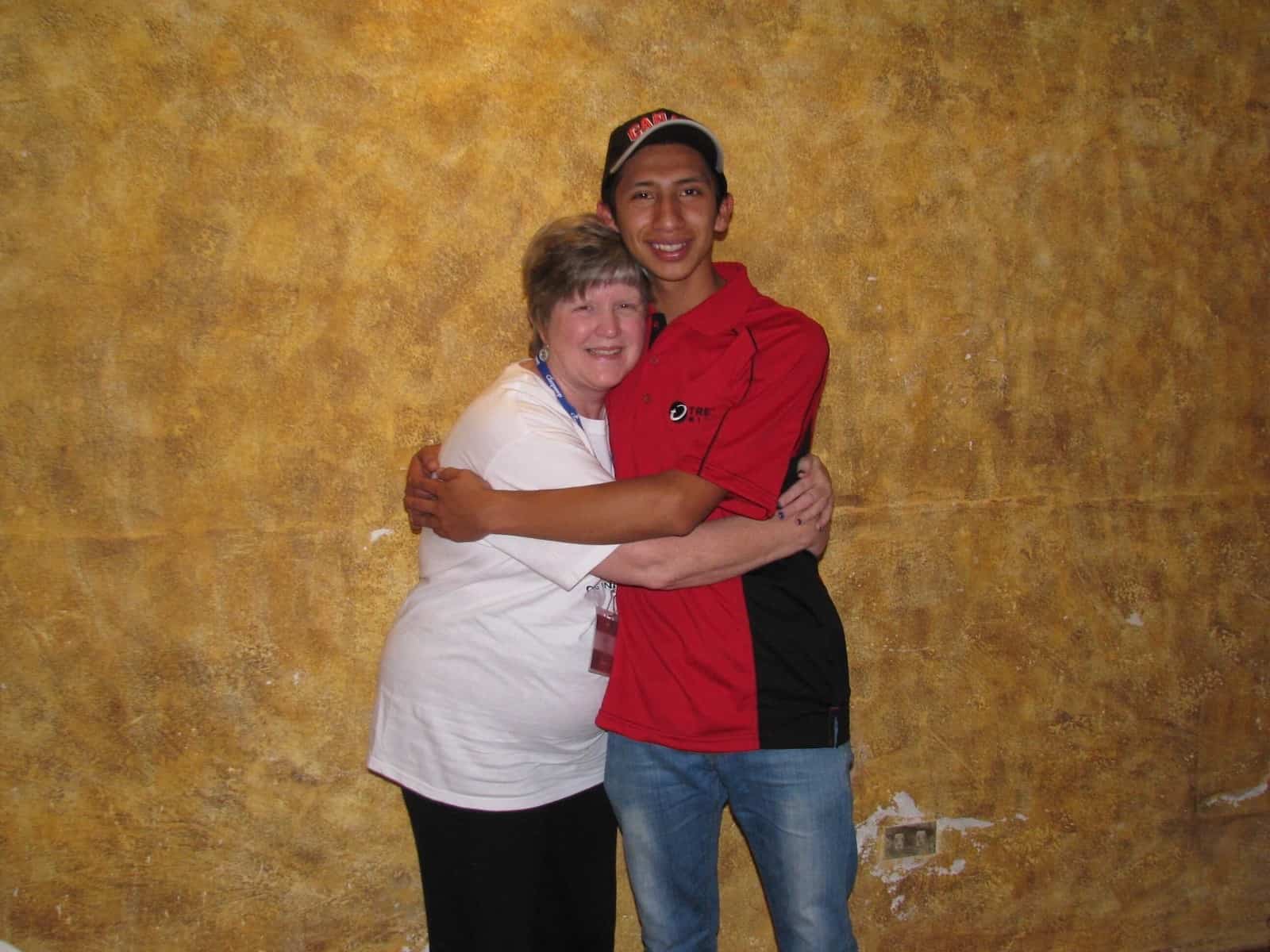 A woman in a white shirt and a young man in a red shirt hug one another tightly.