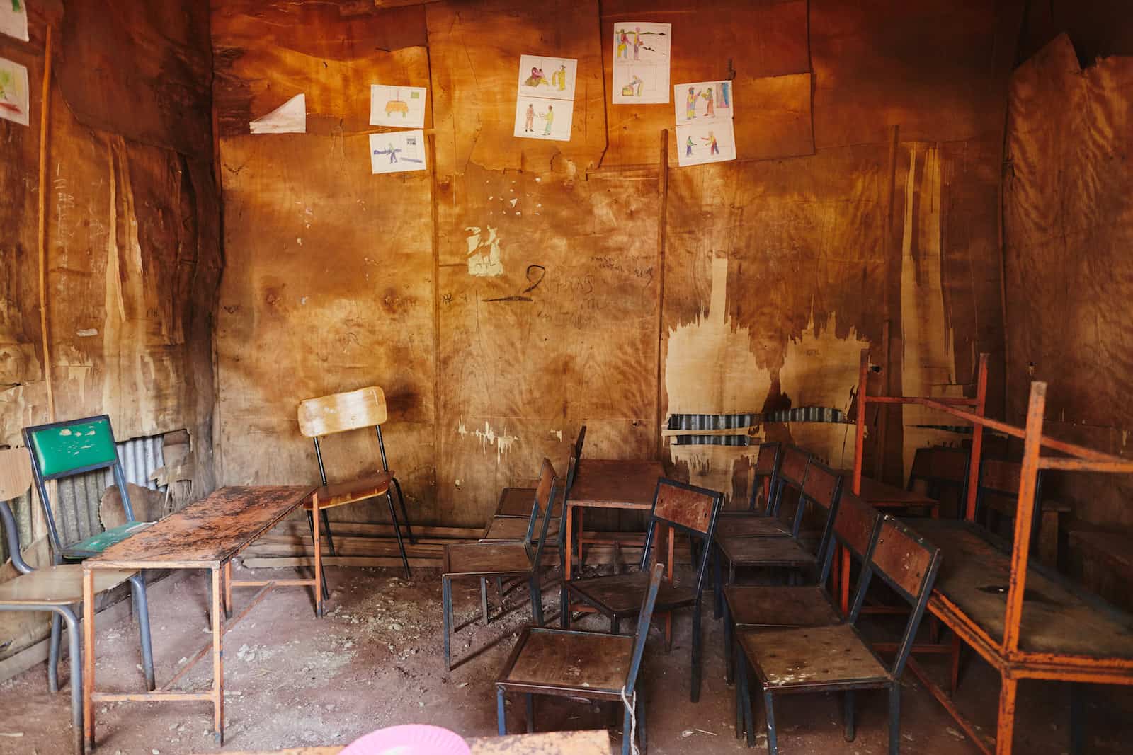 A classroom with dirt floors, plywood walls and cramped chairs.