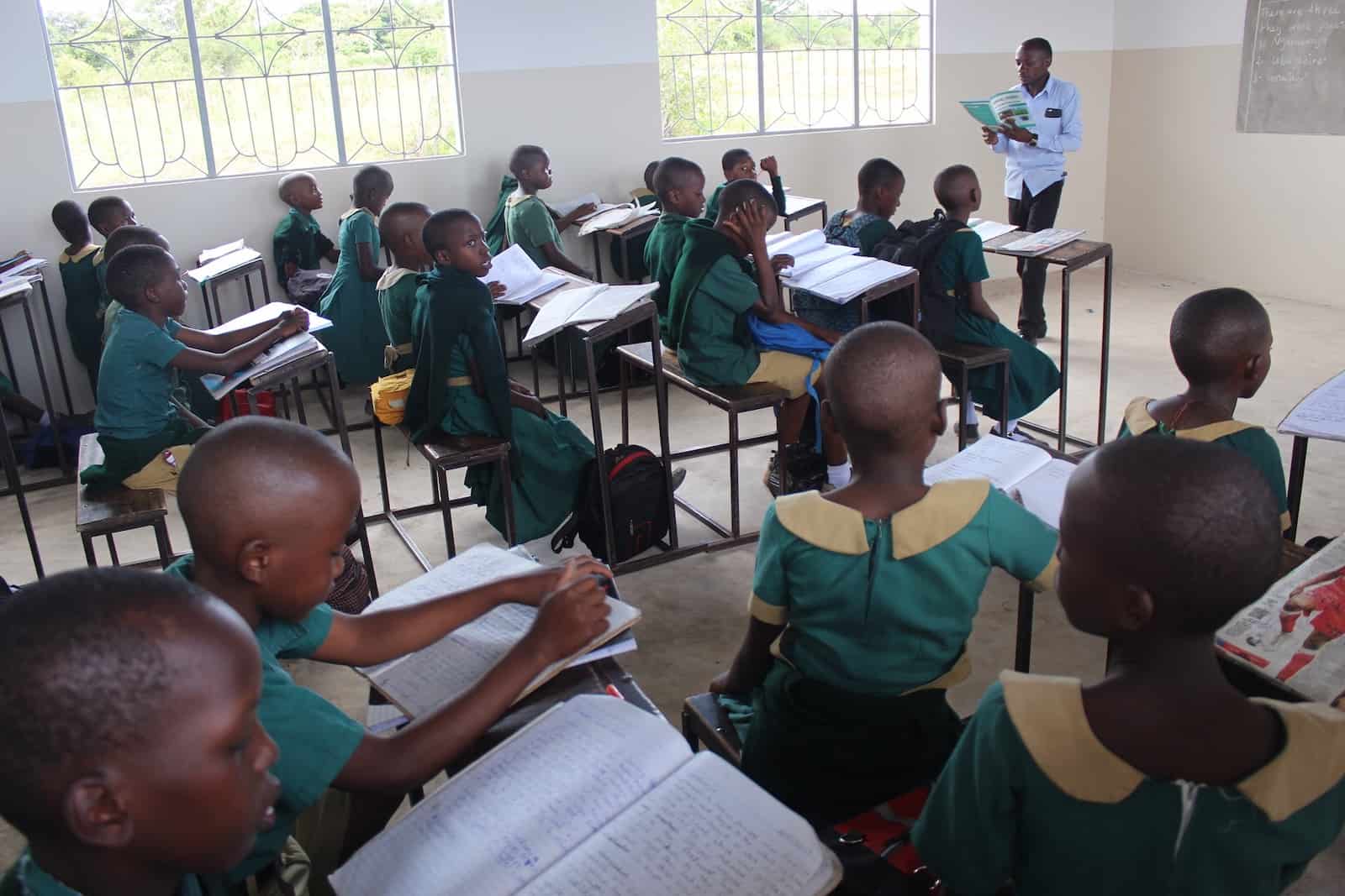 Students sit inside a large classroom with desks and chairs, listening to a teacher. 