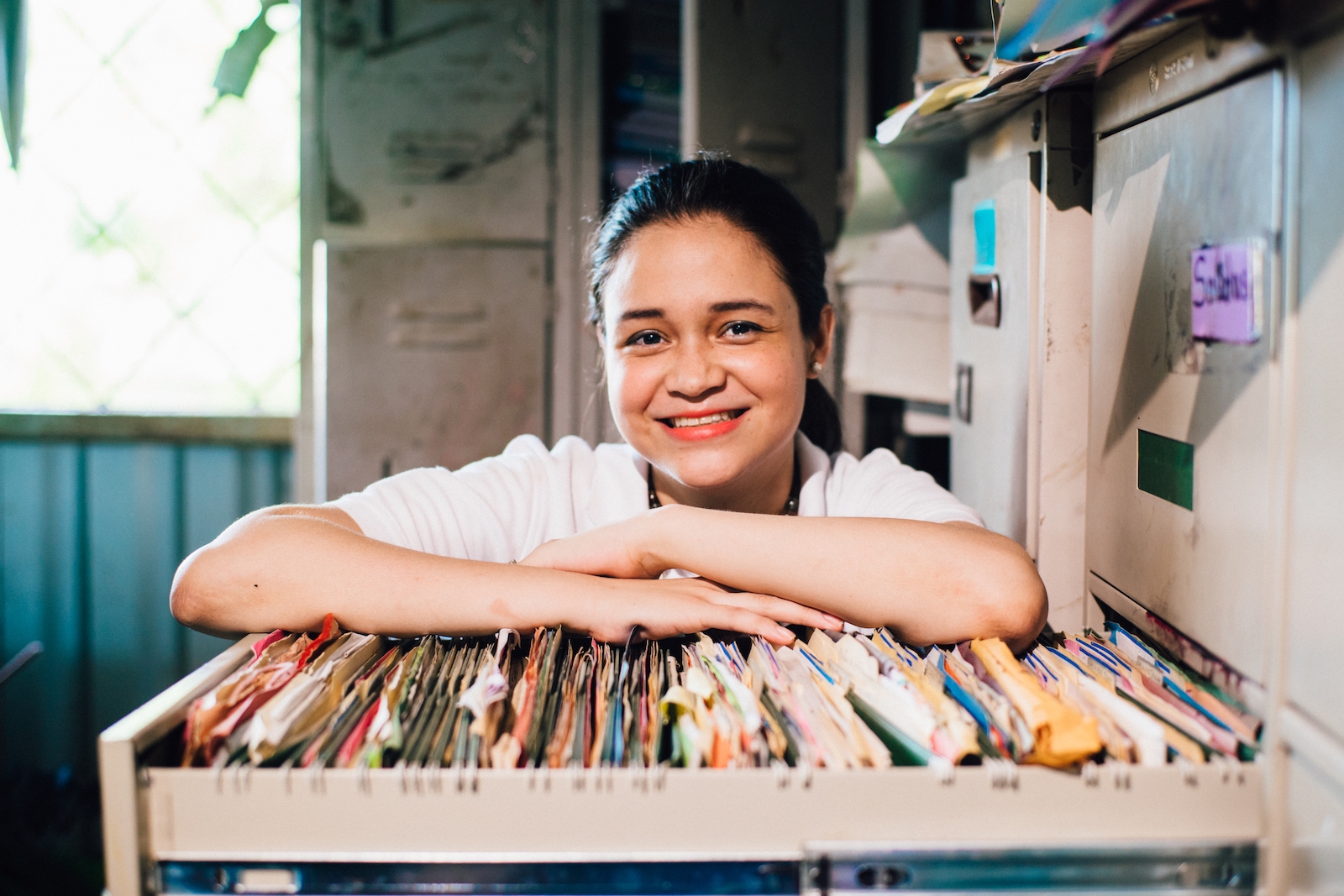A woman leans on a drawer of file folders, smiling, showing the relation between poverty and research.
