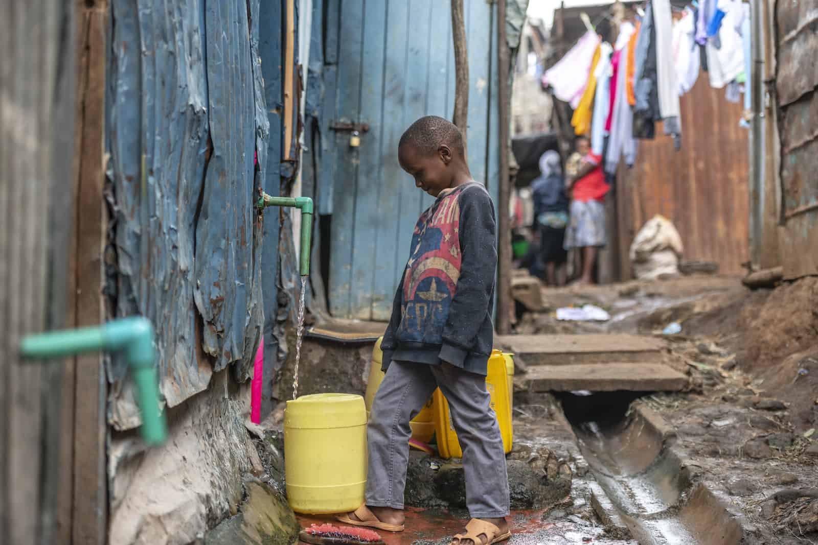 A boy stands in an alley filling a water jug at a tap.