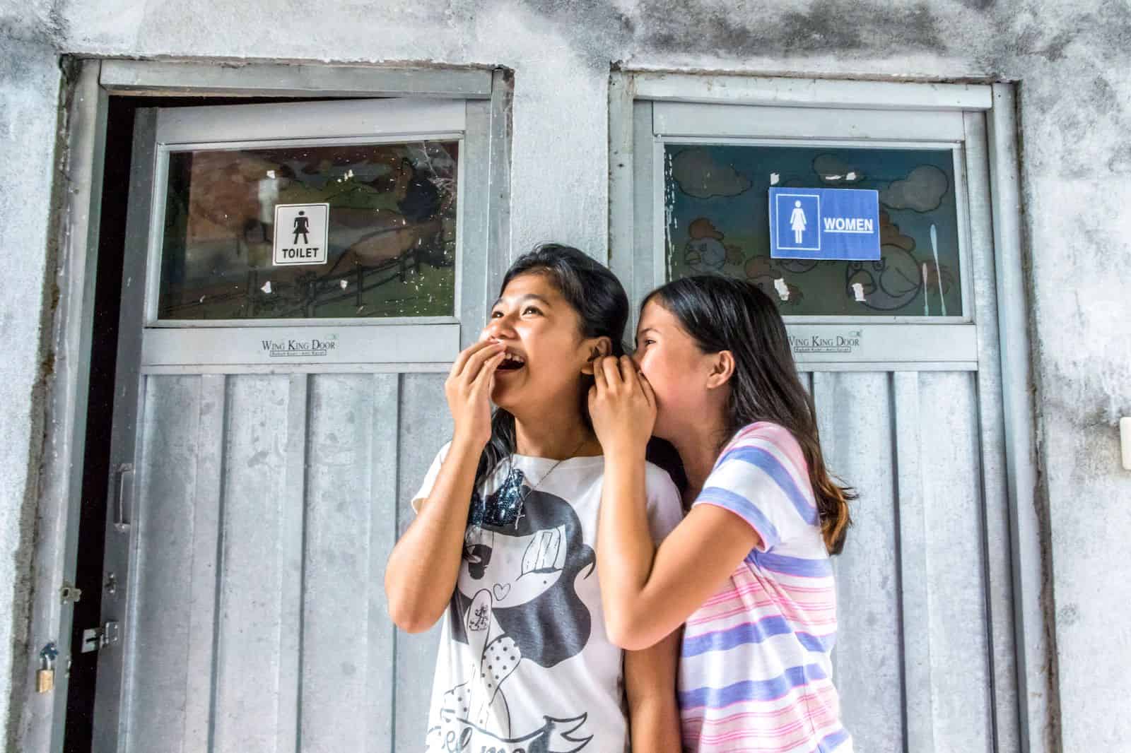 Two girls stand outside a bathroom, giggling.