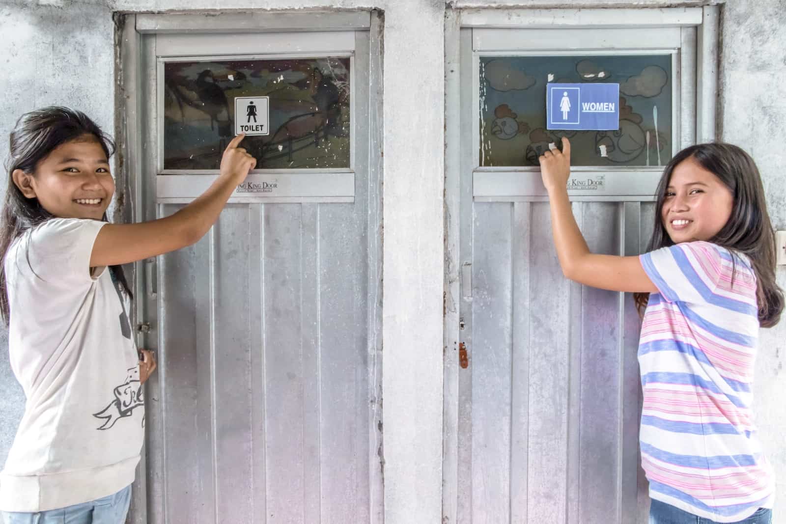 Two girls stand outside bathrooms, pointing at the "women's" signs.