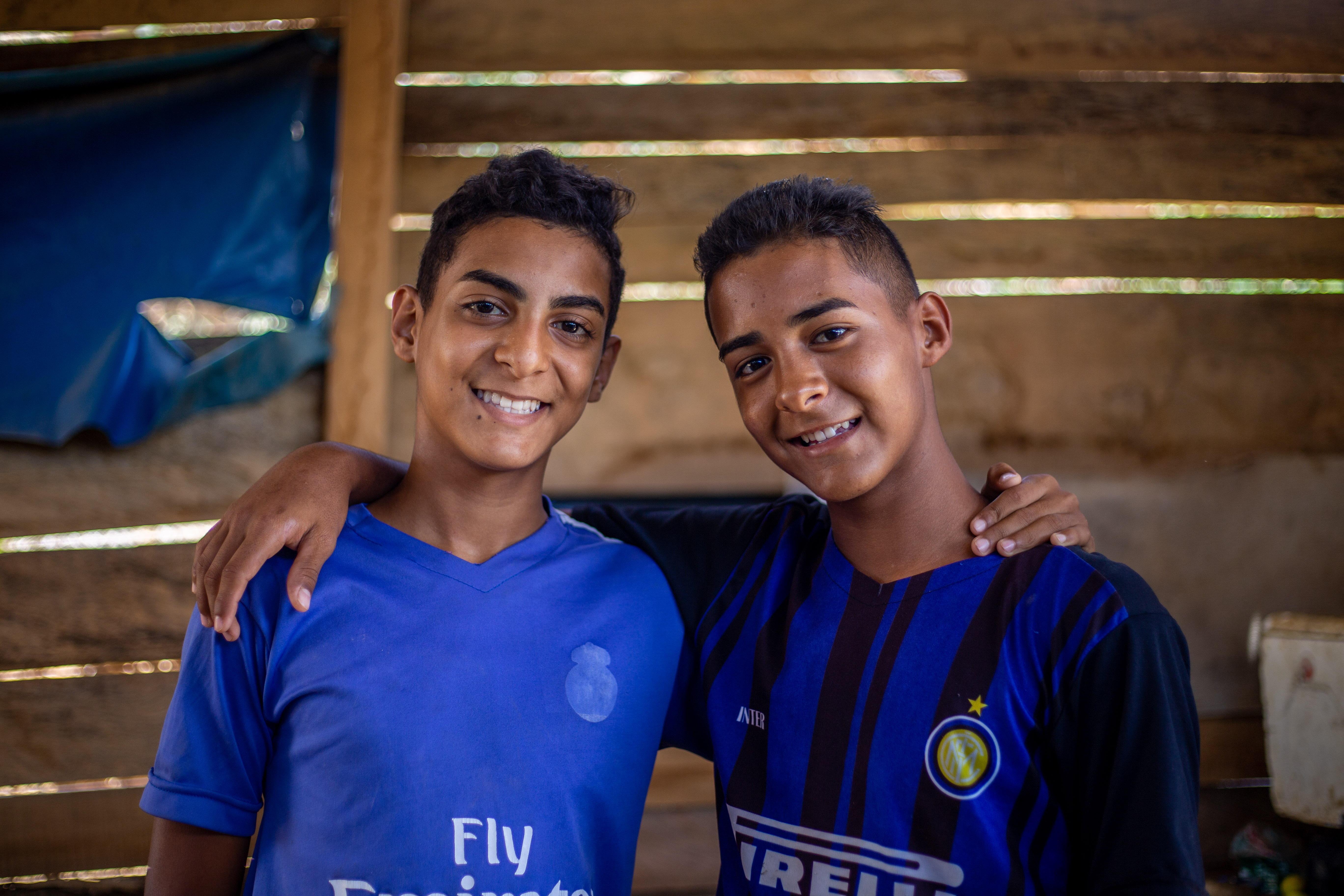 Two teen boys wearing blue and black shirts have their arms around each other, smiling at the camera. 