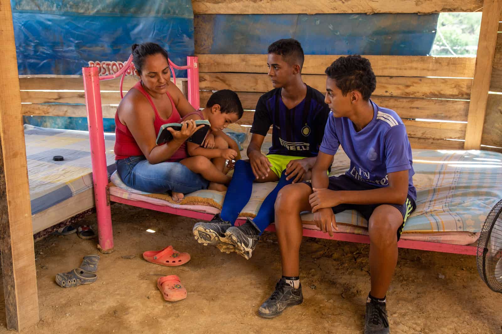 A woman and three boys sit on a bed reading the Bible, in a house with wooden slat walls and dirt floors.
